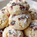 Soft white chocolate cranberry cookies coated in snowy powdered sugar, arranged on a white plate with gentle Christmas lighting.